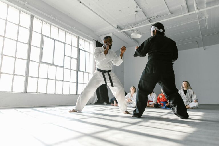 Two martial artists sparring in a dojo with a group watching attentively.